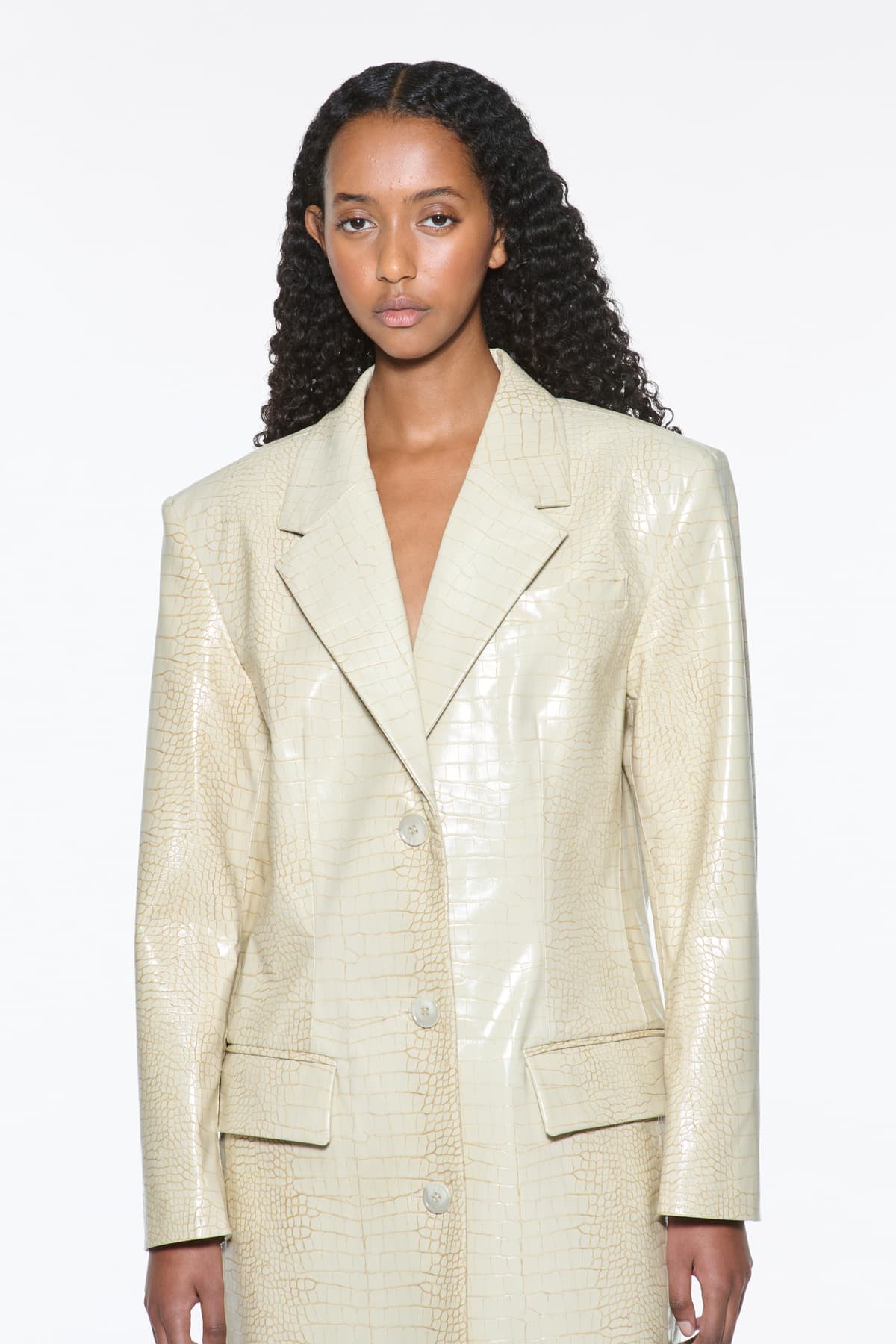 Close-up view of the Nicki Coat in Seashell, is a faux leather trench coat with a shiny croco pattern. The color is a creamy white tone.