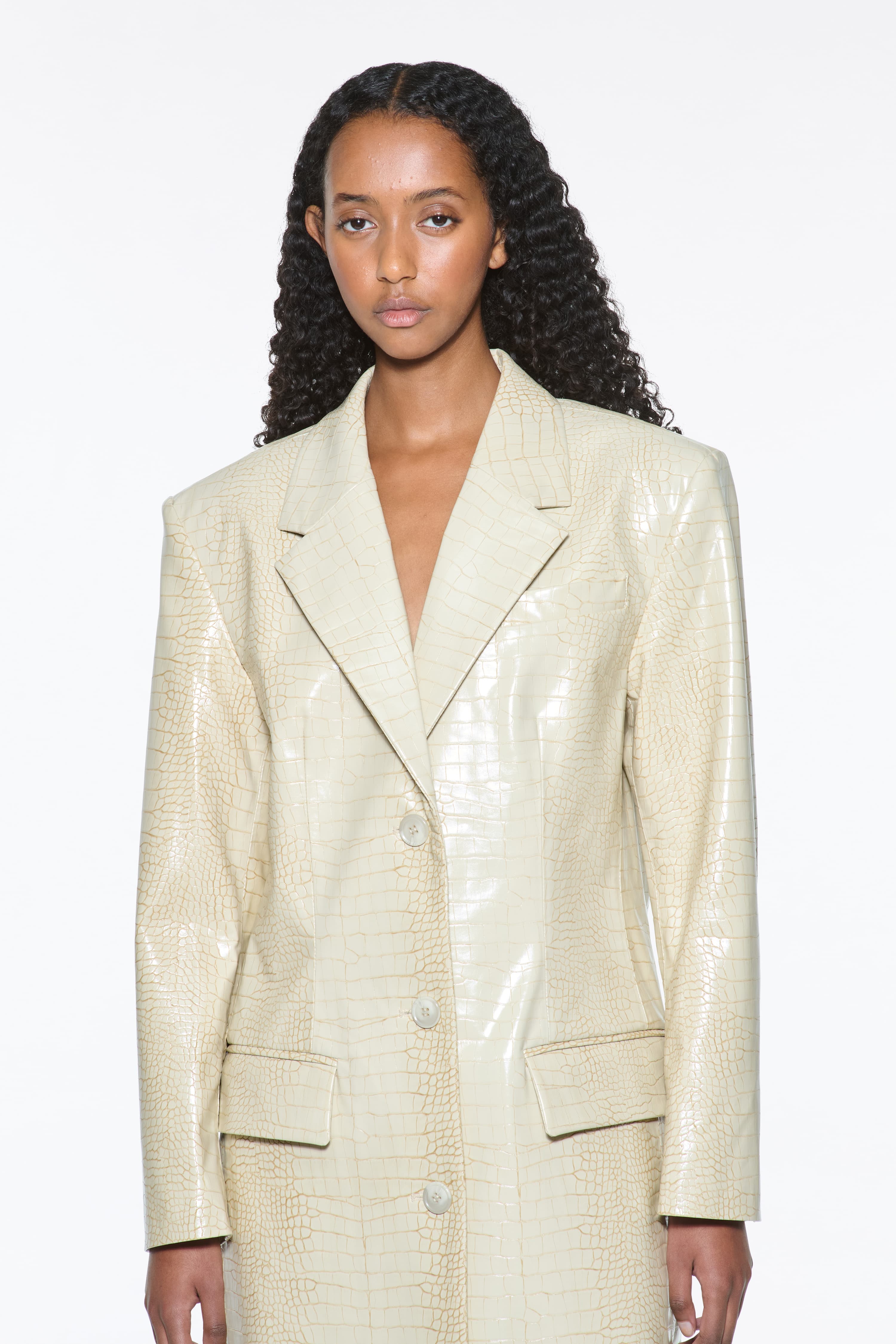 Close-up view of the Nicki Coat in Seashell, is a faux leather trench coat with a shiny croco pattern. The color is a creamy white tone.
