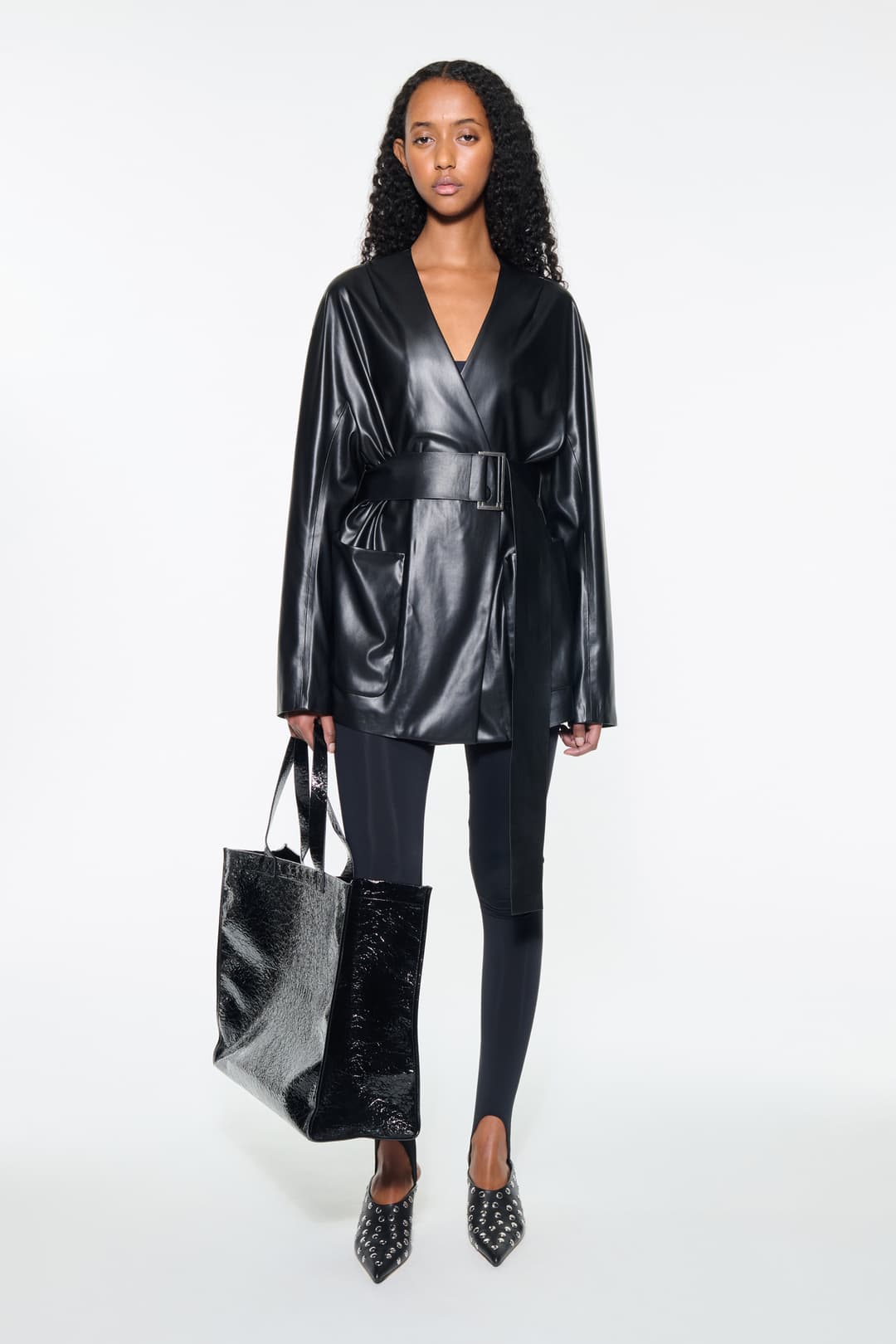 Front view of the Noria Jacket in Black, is a faux leather jacket with a wrap design and a belted waist.