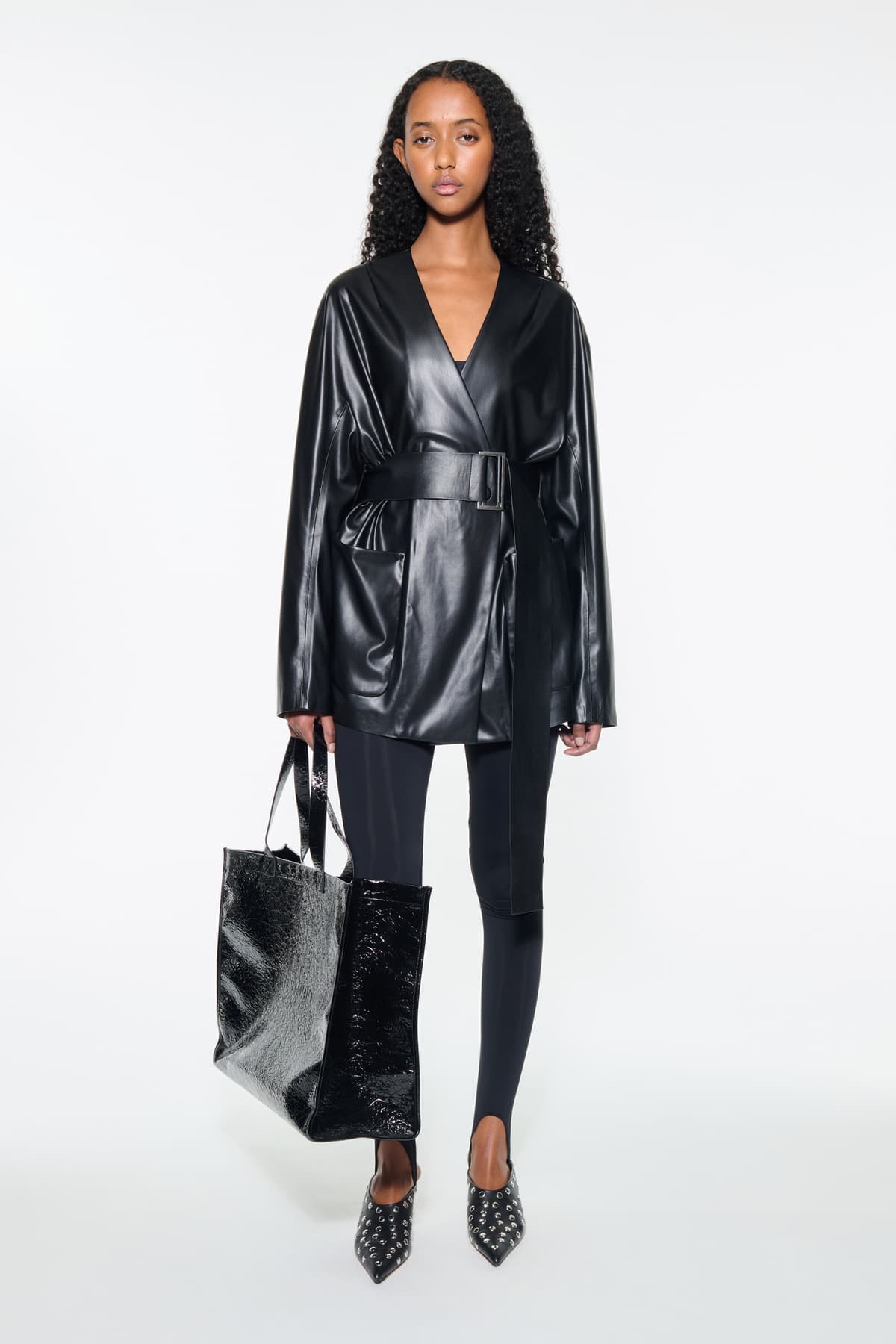 Front view of the Noria Jacket in Black, is a faux leather jacket with a wrap design and a belted waist.
