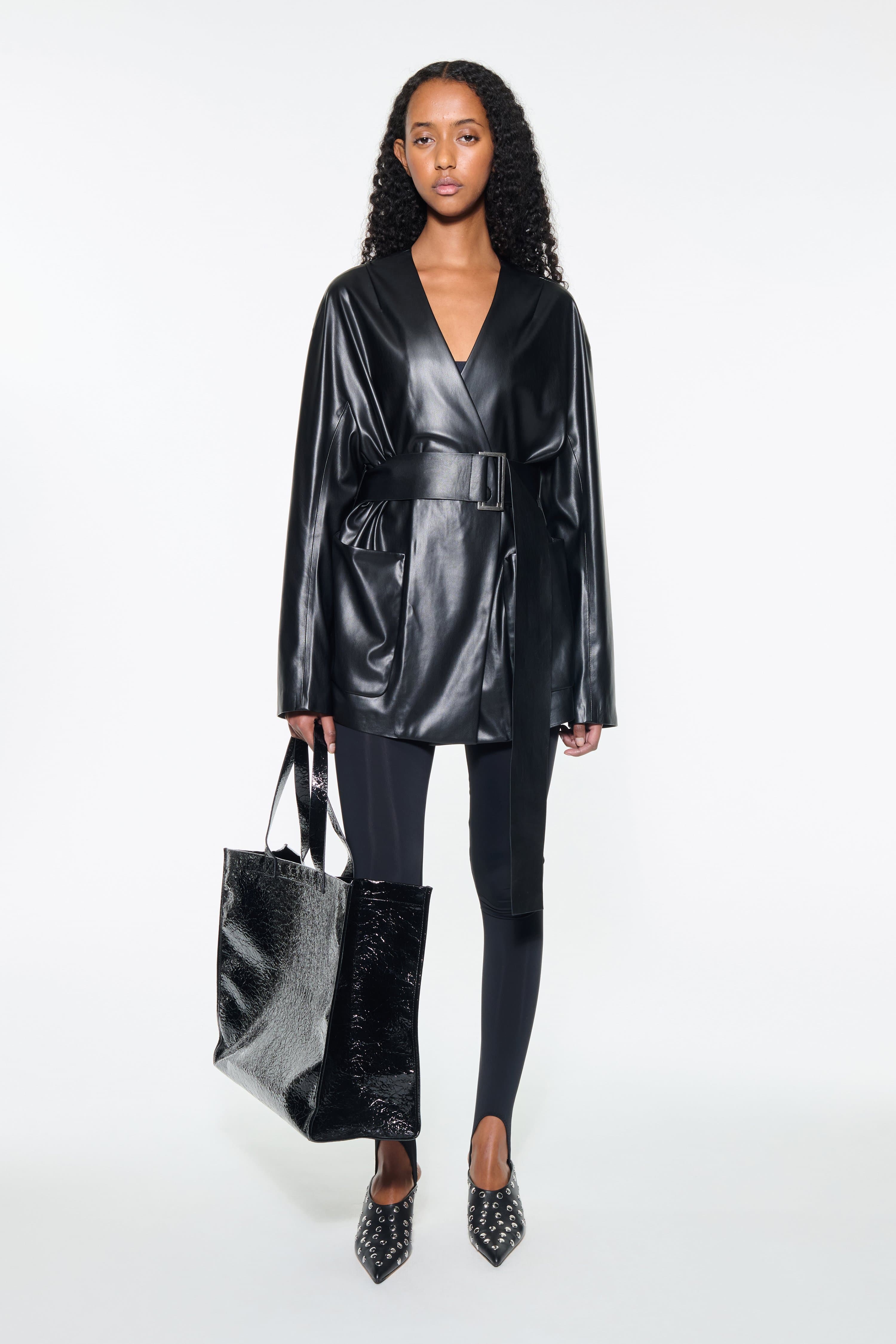 Front view of the Noria Jacket in Black, is a faux leather jacket with a wrap design and a belted waist.