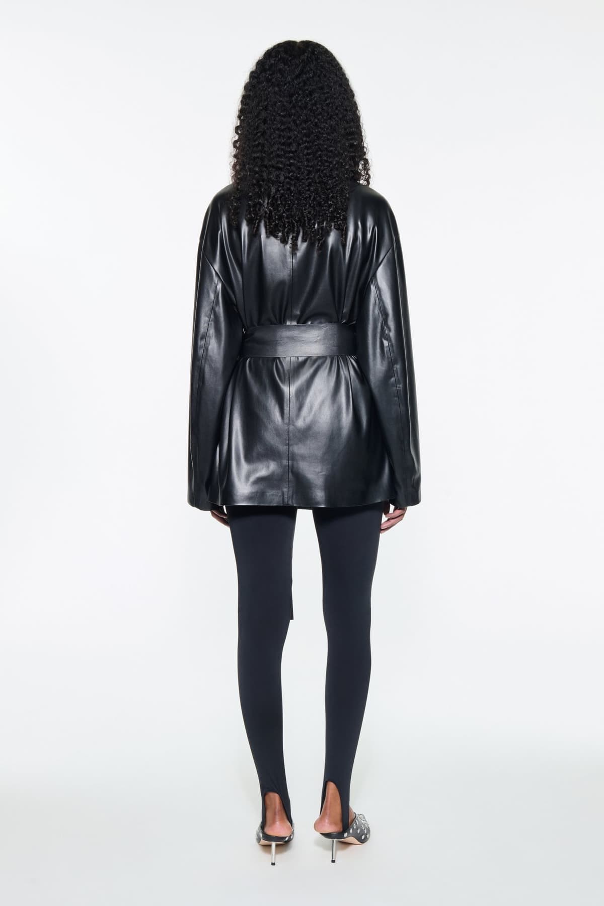 Back view of the Noria Jacket in Black, is a faux leather jacket with a wrap design and a belted waist.