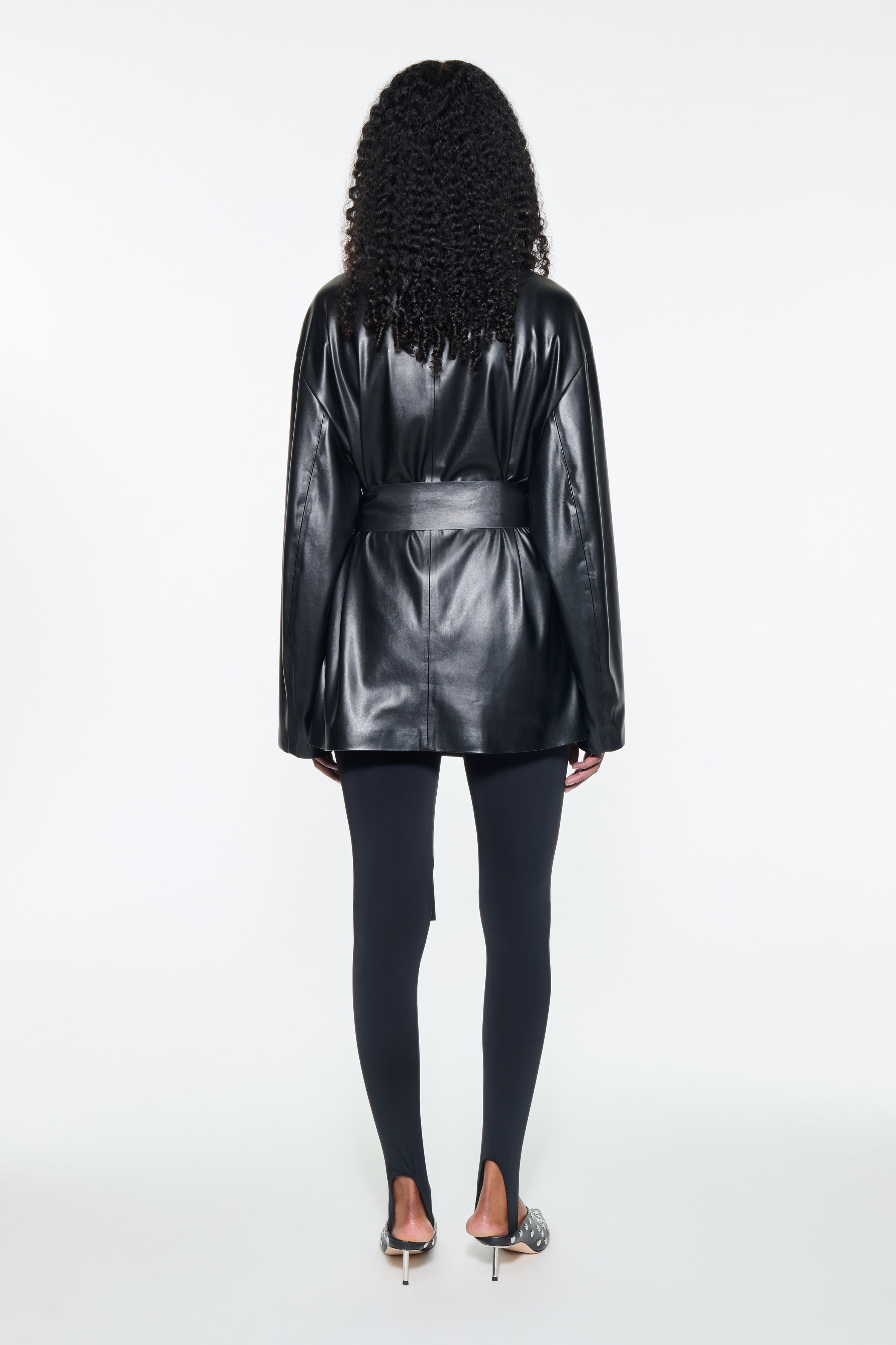 Back view of the Noria Jacket in Black, is a faux leather jacket with a wrap design and a belted waist.