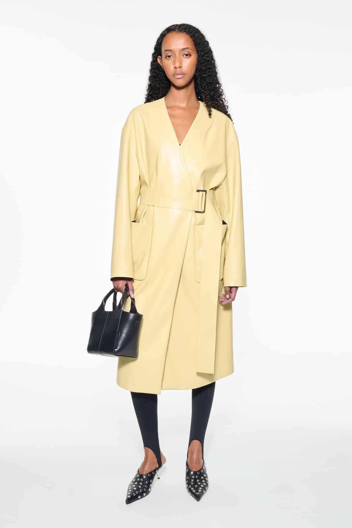 Front view of the Valeria Coat in Pina Colada, is a faux leather coat with a wrap design and a belted waist. The color is a pale yellow tone.