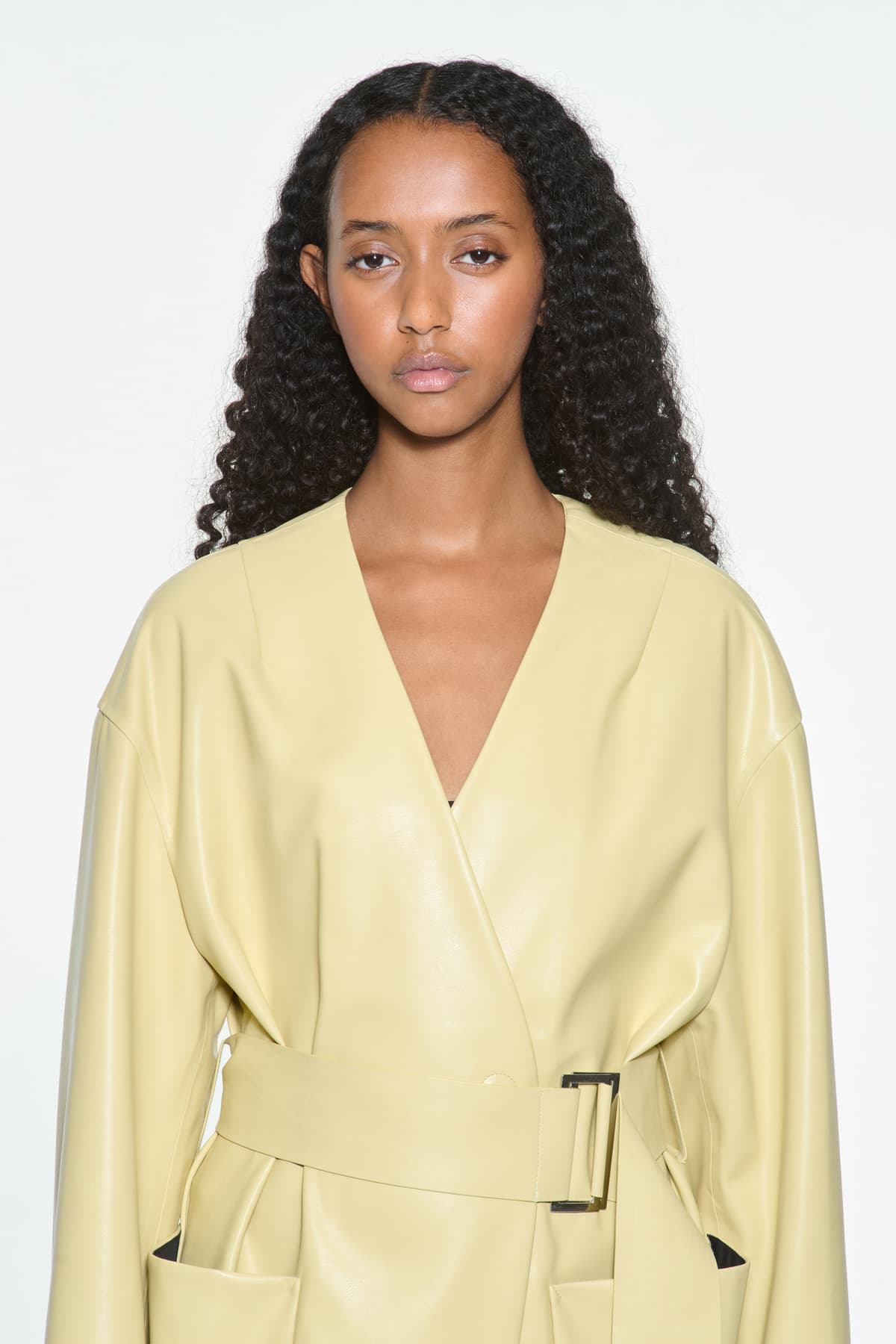 Close-up view of the Valeria Coat in Pina Colada, is a faux leather coat with a wrap design and a belted waist. The color is a pale yellow tone.