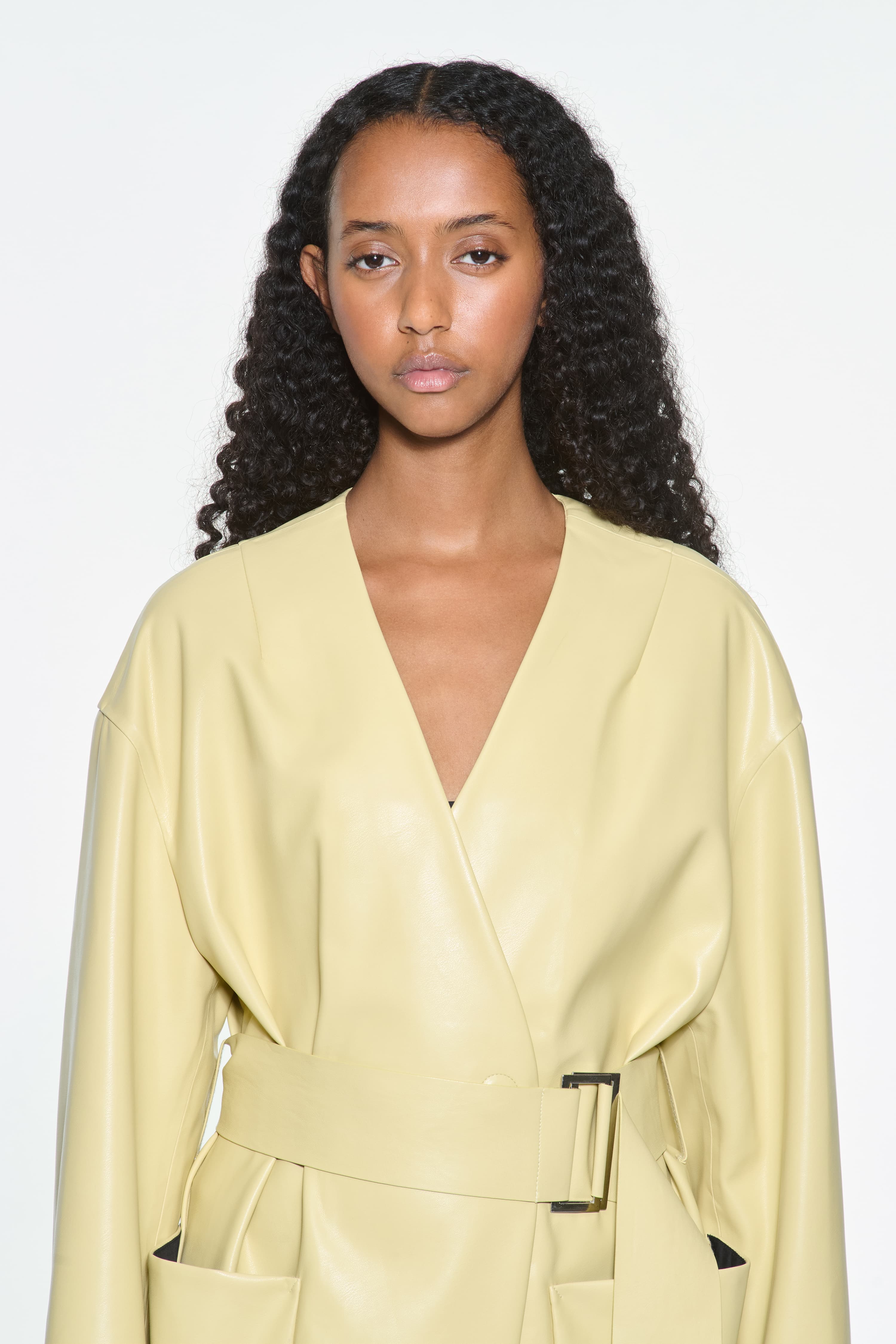 Close-up view of the Valeria Coat in Pina Colada, is a faux leather coat with a wrap design and a belted waist. The color is a pale yellow tone.
