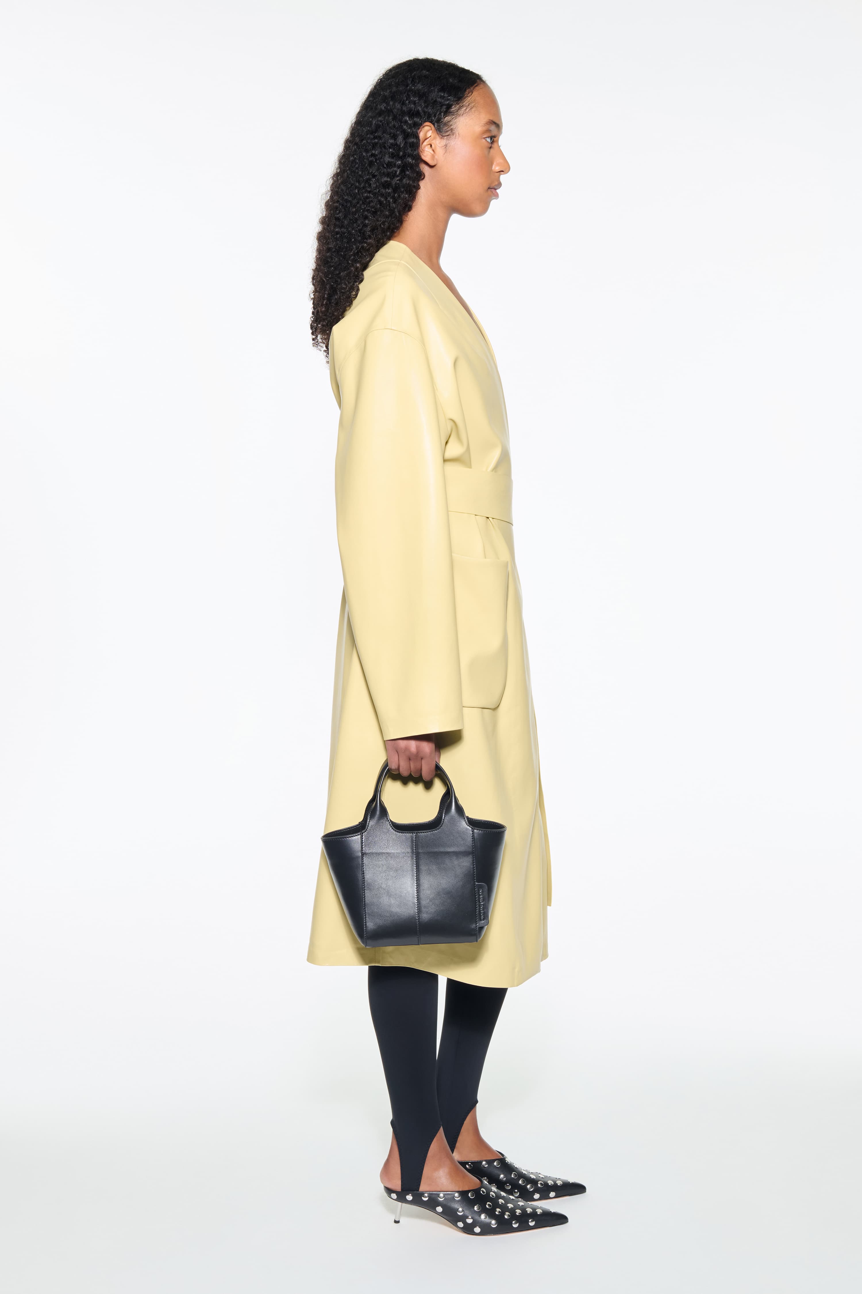 Side view of the Valeria Coat in Pina Colada, is a faux leather coat with a wrap design and a belted waist. The color is a pale yellow tone.